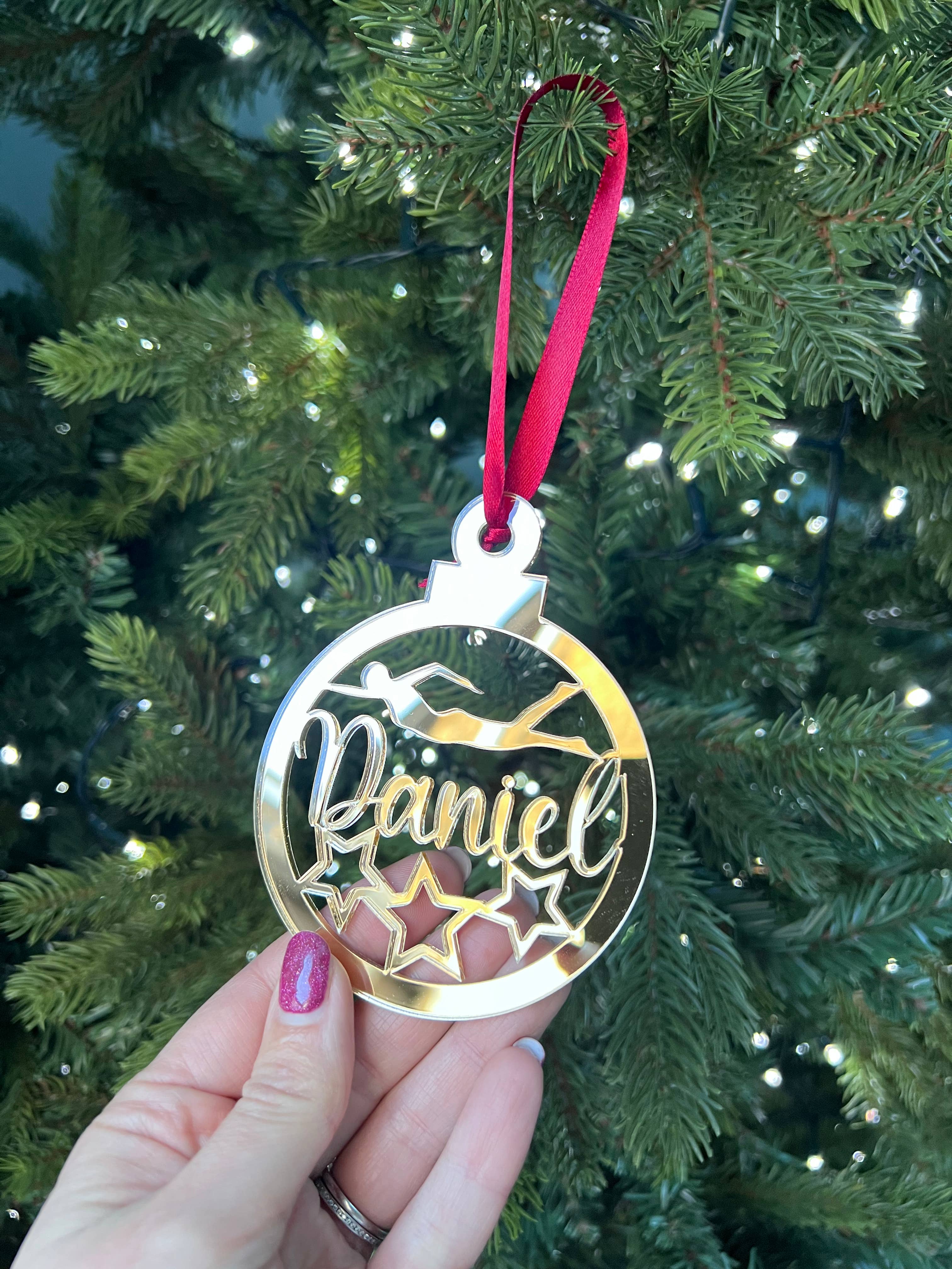 A hand holding a gold-colored Christmas tree decoration with the name 'Daniel' engraved on it, against a blurred background of a Christmas tree with lights.